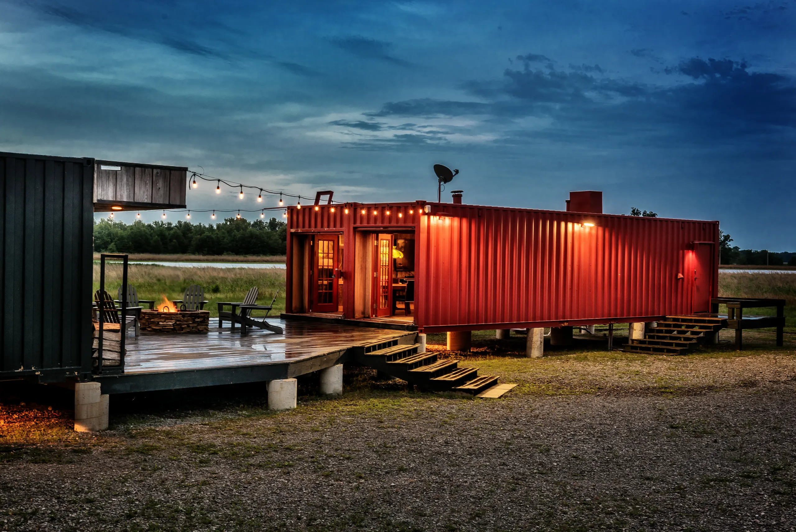 The red shipping container lodge and its deck at dusk, string lights overhead